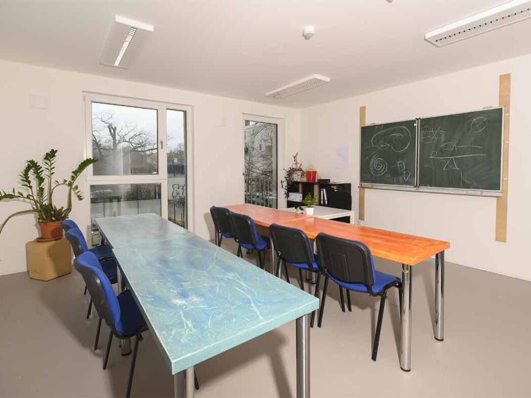 Innenansicht degewo-Neubau Gotenburger Straße 2: Klassenzimmer der Jugendberufshilfe mit zwei bestuhlten Tischreihen und Wandtafel.