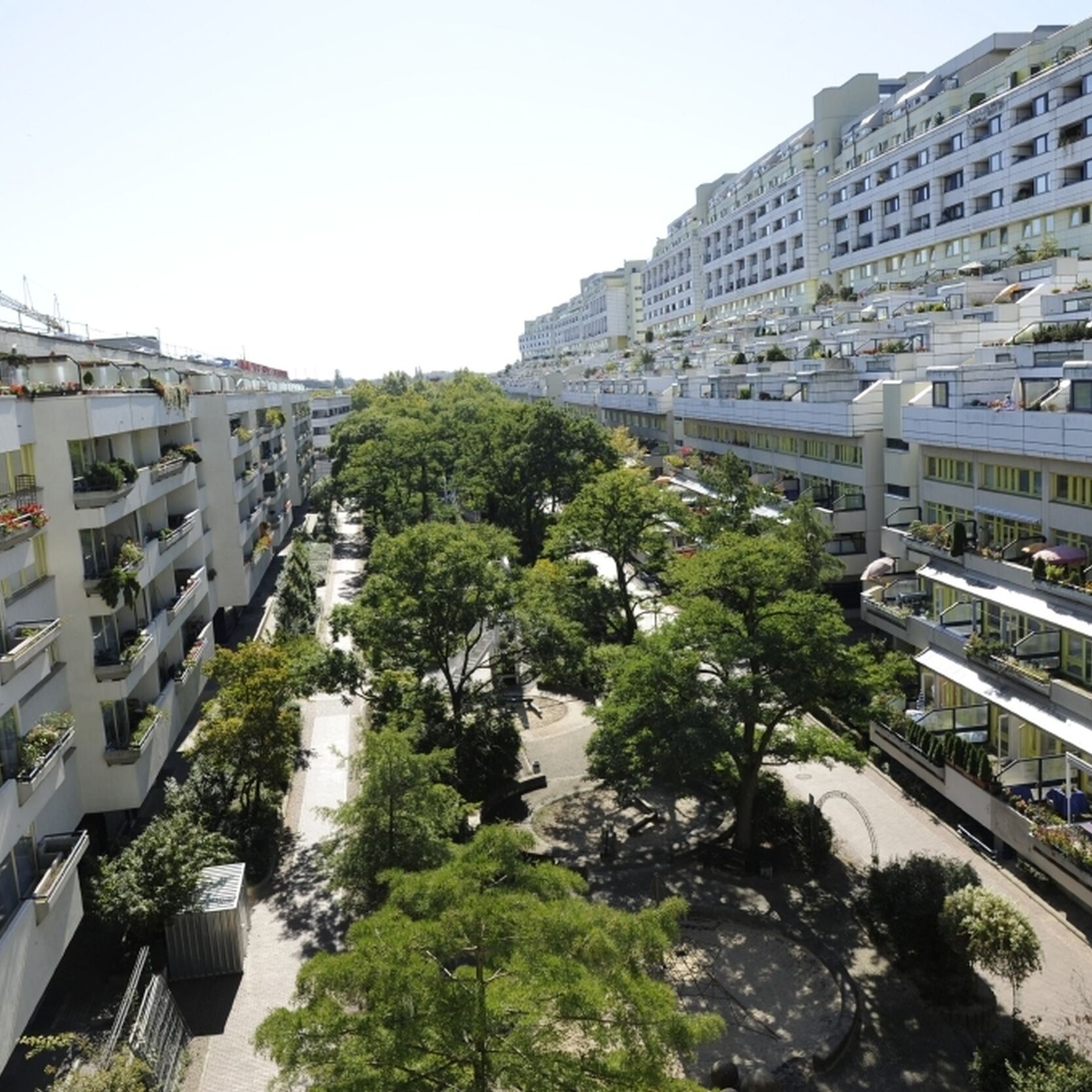 Blick von oben auf Gebäudezeilen links und rechts sowie hofseitige Mittelinsel mit schattenspendendem Baumbestand.