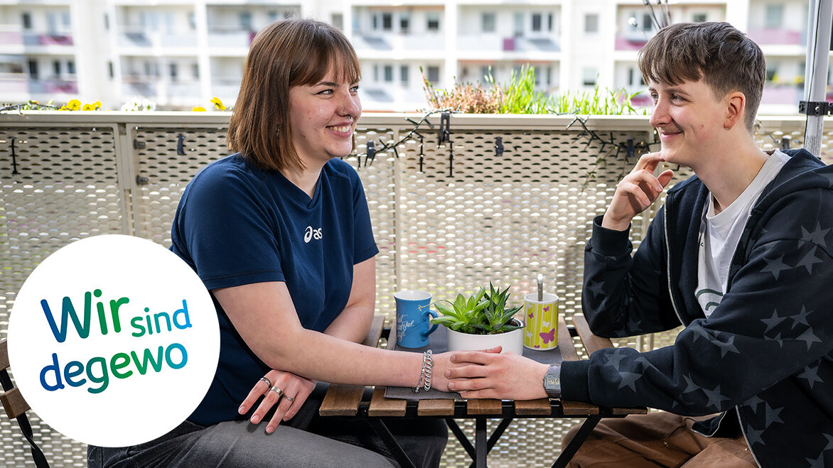 Ein junges Paar sitzt auf dem Balkon auf Stühlen gemeinsam an einem Tisch. Sie schauen sich in die Augen und halten Händchen. Einige Pflanzen zieren den Balkon.