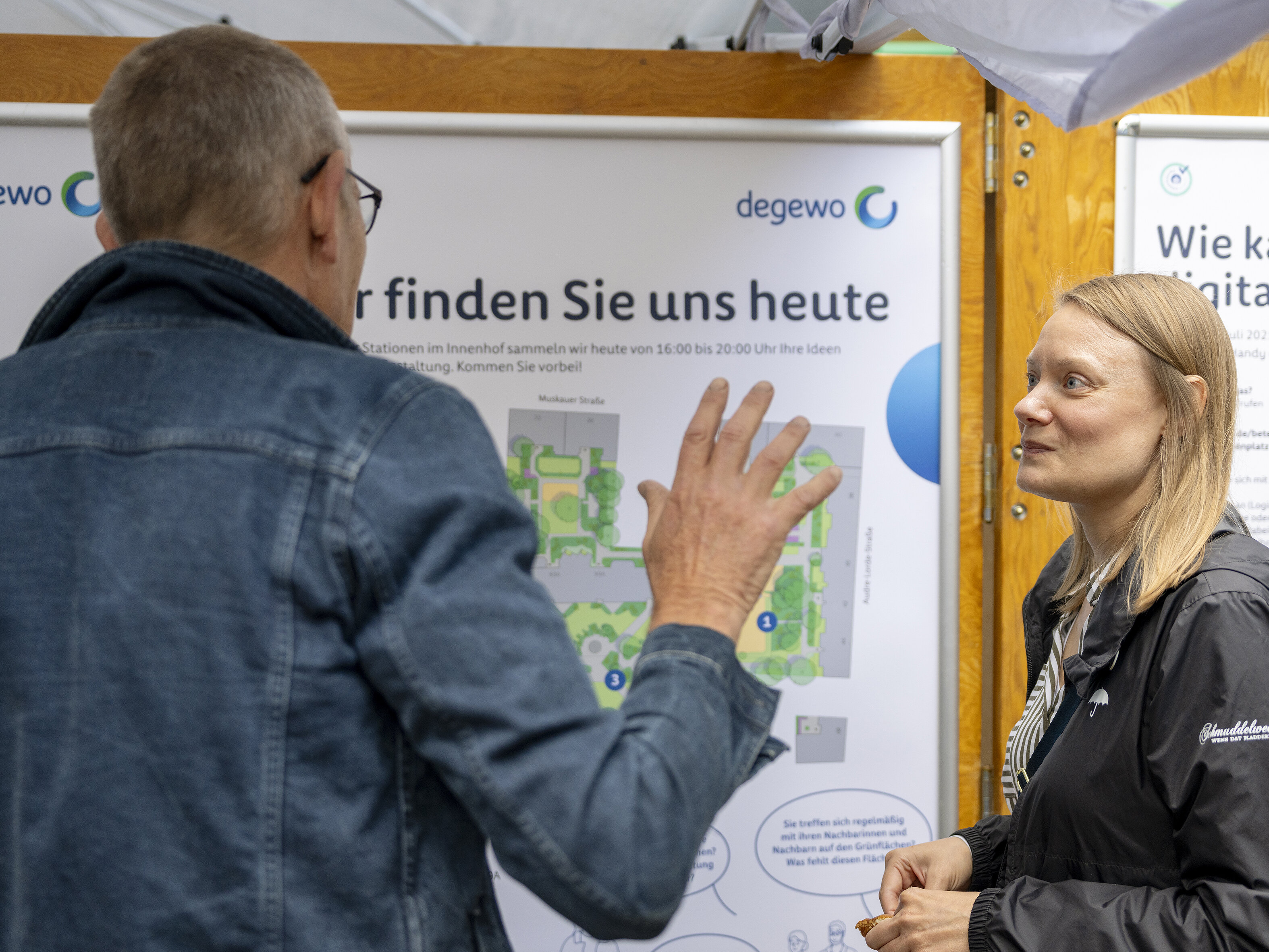 Austausch zwischen einem Bewohner und einer Mitarbeiterin vor einer Informationswand mit Lageplan und Hinweisen zur Beteiligung.