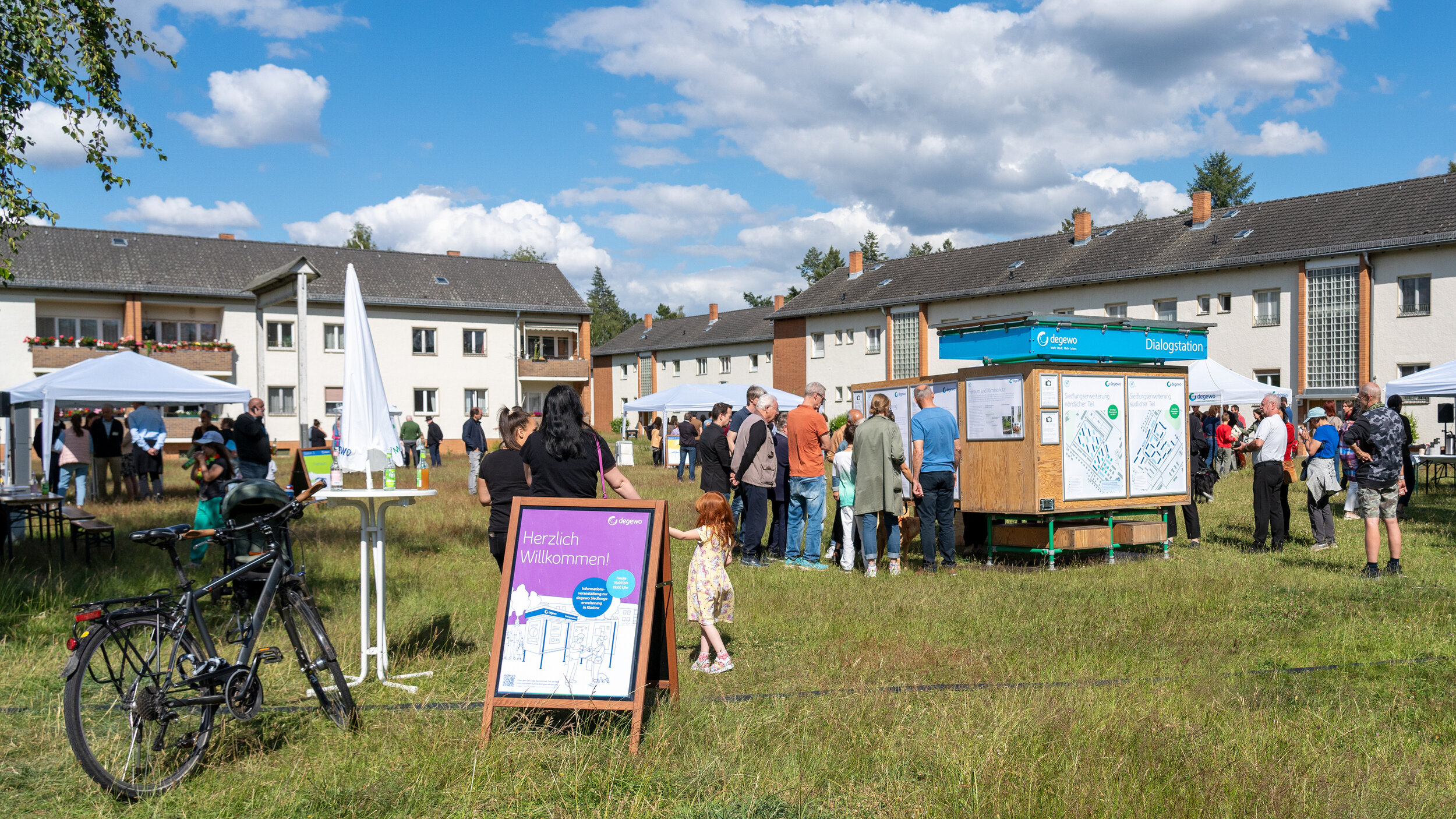 Menschen stehen auf einer Wiese im Freien, auf dieser sind verschiedene Infostände verteilt. Im Hintergrund befinden sich Wohnhäuser.
