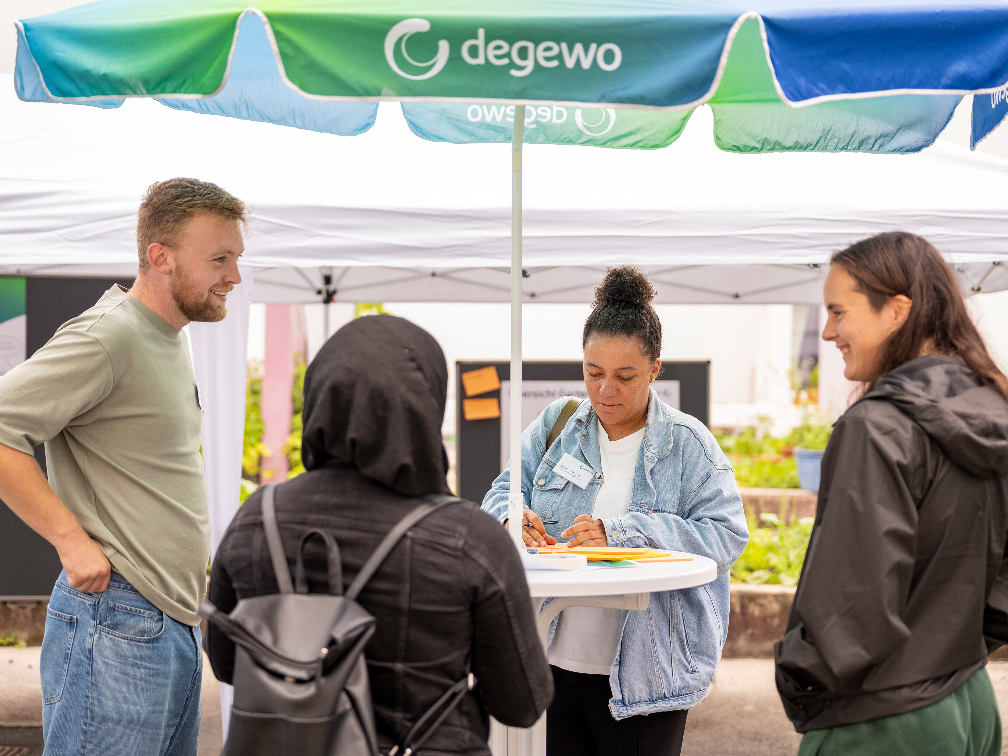 Austausch am Stehtisch unter einem degewo-Sonnenschirm: Eine Mitarbeiterin notiert Anmerkungen, während mehrere Teilnehmende ihre Ideen zur Hofgestaltung teilen.