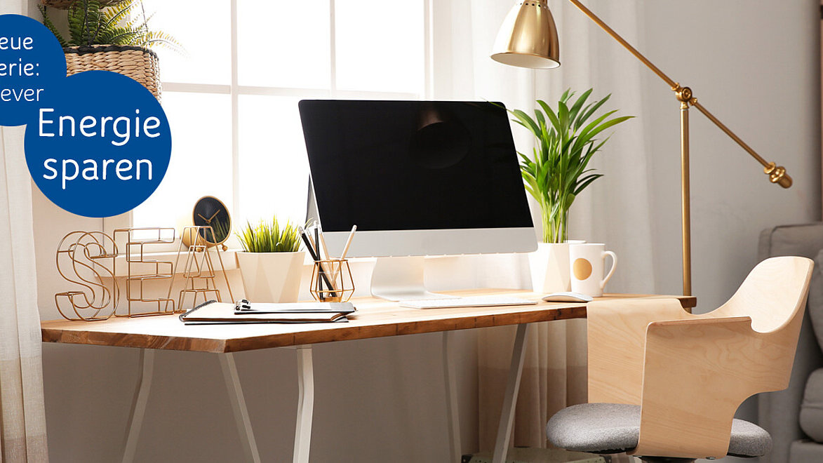 Auf einem Schreibtisch am Fenster sind Laptop und ein großer Bildschirm platziert. Auf der Tischplatte liegt weiteres Arbeitsmaterial, die Sonne scheint durch die große Fensterfront hinein.
