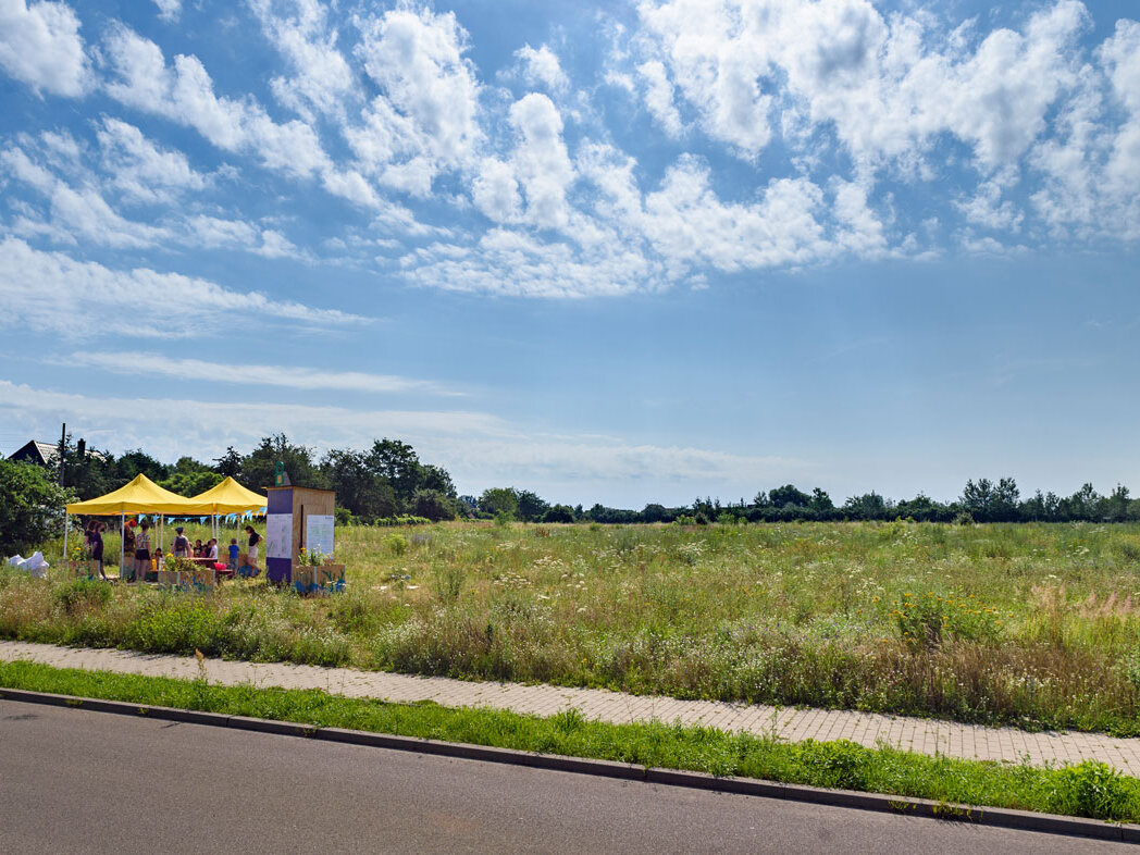 Weitläufige Grünfläche mit einem Beteiligungsstand unter gelben Zelten. Mehrere Menschen informieren sich über ein Projekt, im Hintergrund ist offene Landschaft zu sehen.