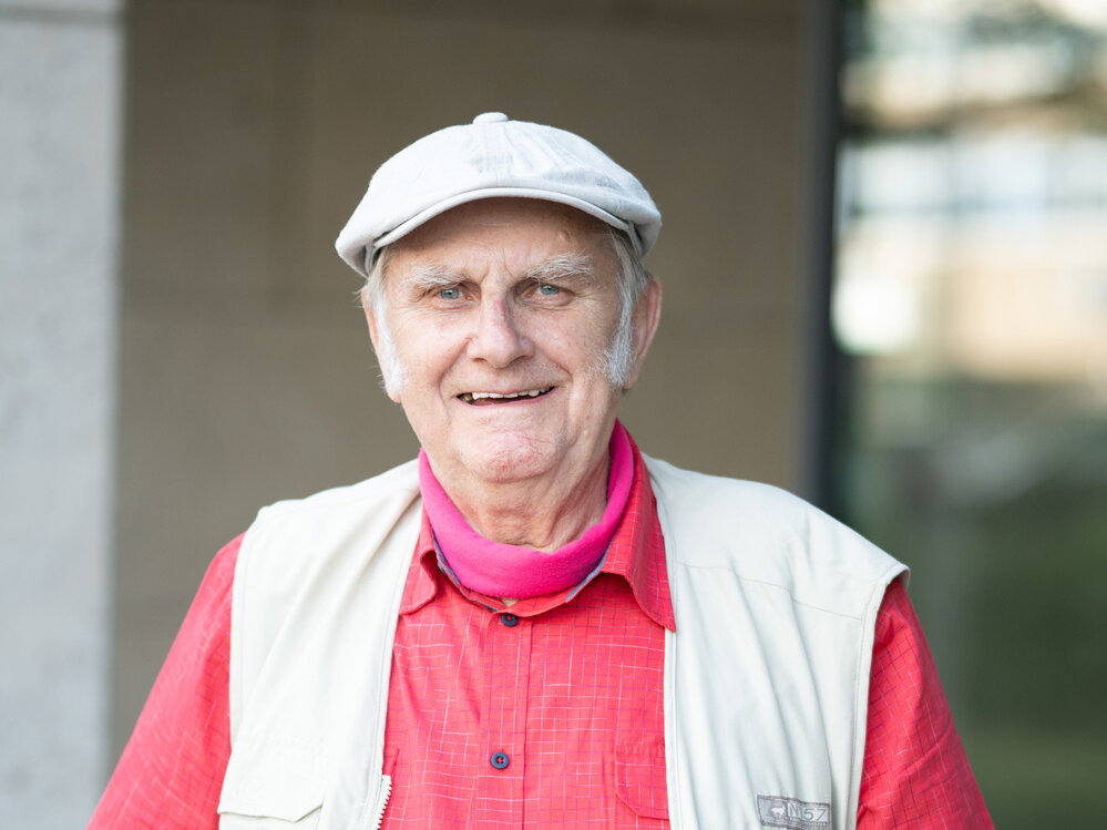 Älterer Mann mit heller Weste, rotem Hemd und Schiebermütze steht im Freien vor einem Gebäude und lächelt leicht; der Hintergrund ist unscharf und zurückhaltend.