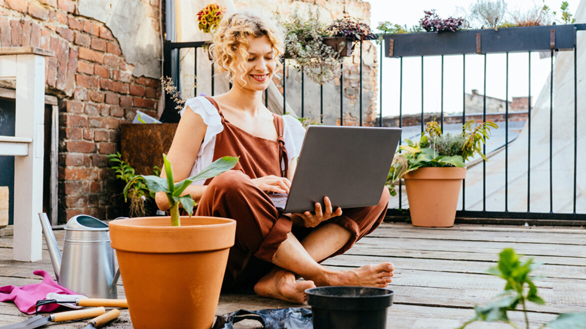 Eine blonde Frau sitzt auf einem Balkon im Schneidersitz und hat einen Laptop in der Hand. Um sie herum stehen Blumentöpfe und Werkzeuge zum Gärtnern.
