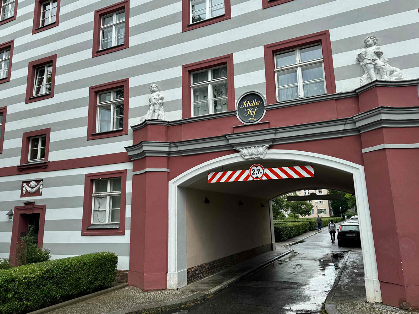 Ein weiß-grau gestreiftes Gebäude mit einer großen Toreinfahrt. Über dem Tor ist ein Schmuckelement mit dem Namen "Schillerhof" angebracht.