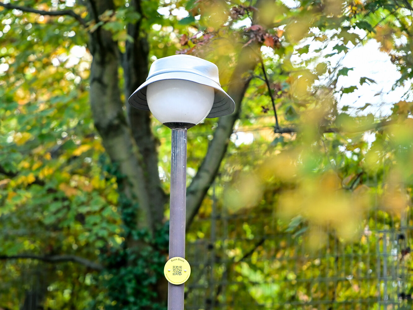 Eine Laterne mit rundem Leuchtenkopf trägt einen hellgrauen Sonnenhut. Im Hintergrund sind herbstliche Bäume zu sehen.