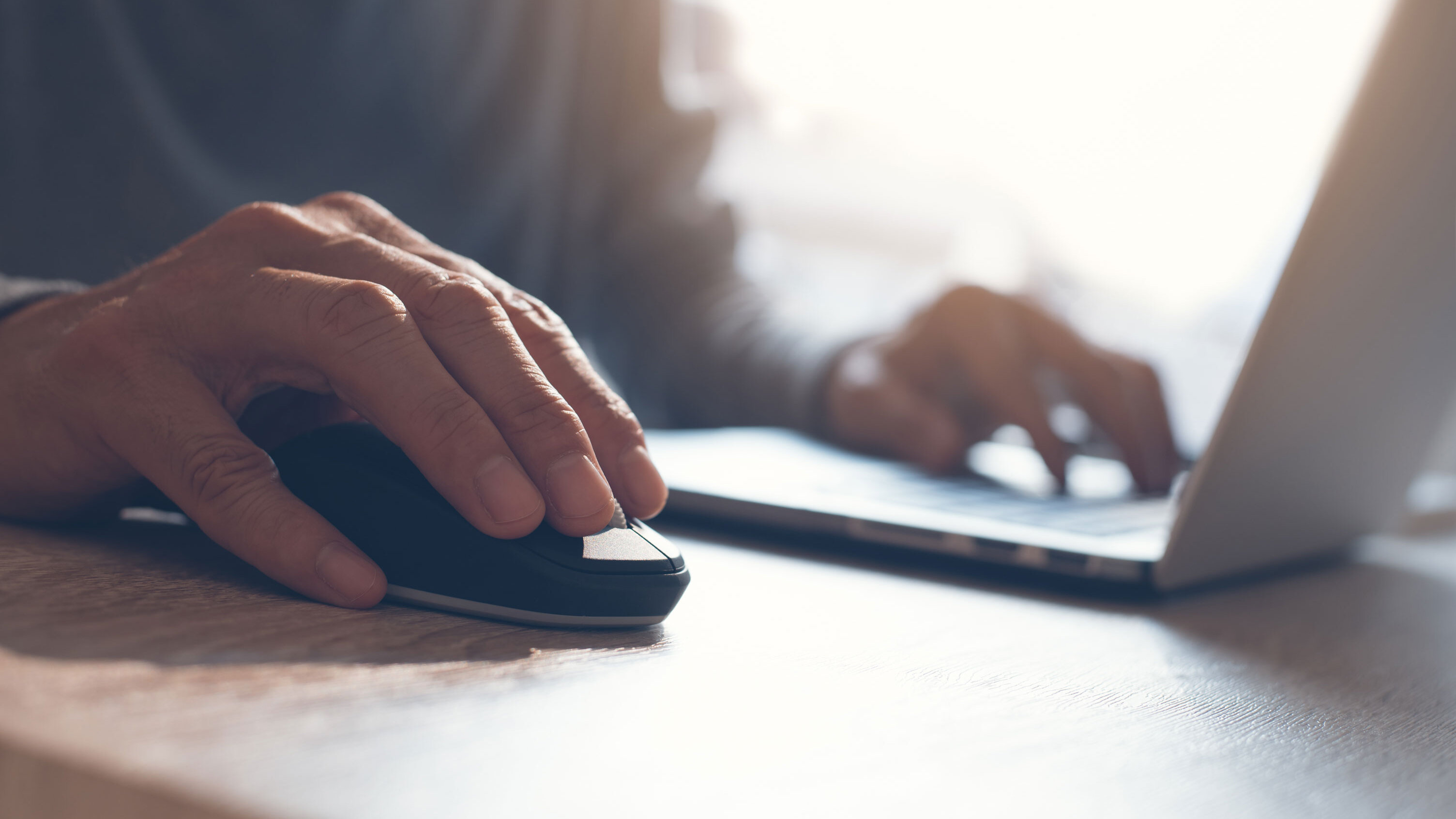 Nahaufnahme einer rechten Hand auf einer schwarzen Computermaus neben einem geöffneten Laptop. Die andere Hand liegt auf der Tastatur. Beide Geräte stehen auf einem Holztisch, der Hintergrund ist unscharf.