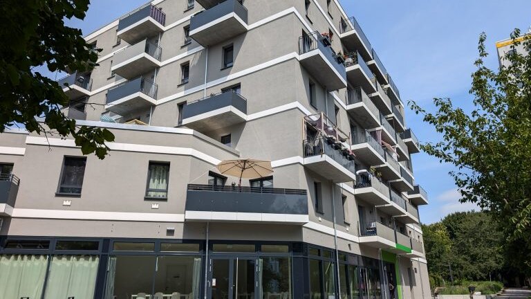 Mehrgeschossiges Wohngebäude mit Balkonen und einem Veranstaltungsraum im Erdgeschoss. Die großen Fensterflächen des Gemeinschaftsraums sind von außen sichtbar, davor befinden sich Gehwege und Grünflächen.