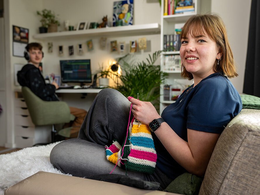 Paar geht Hobbies nach. Mann sitzt am Computer und Frau sitzt auf der Couch und strickt in einem hell eingerichteten Wohnzimmer mit Regalen und Schreibtisch. Beide lächeln in die Kamera.