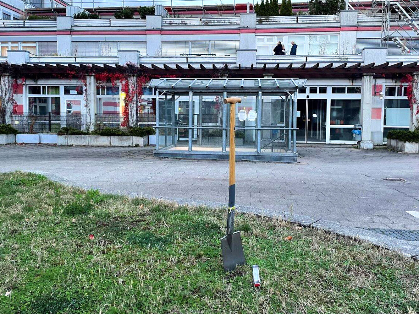 Eine einzelne Schaufel steckt aufrecht in einer Rasenfläche vor einem Gebäudekomplex.