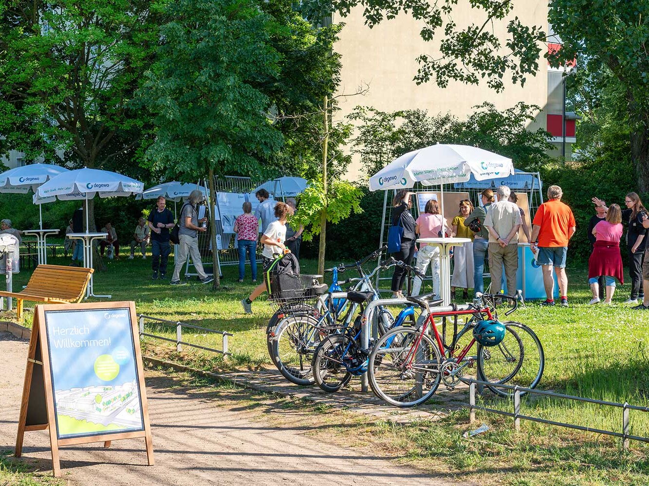 Blick auf eine Infoveranstaltung im Grünen: An mehreren Ständen unter degewo-Sonnenschirmen informieren sich Anwohnende, Fahrräder stehen am Wegesrand, ein Aufsteller heißt die Gäste willkommen.