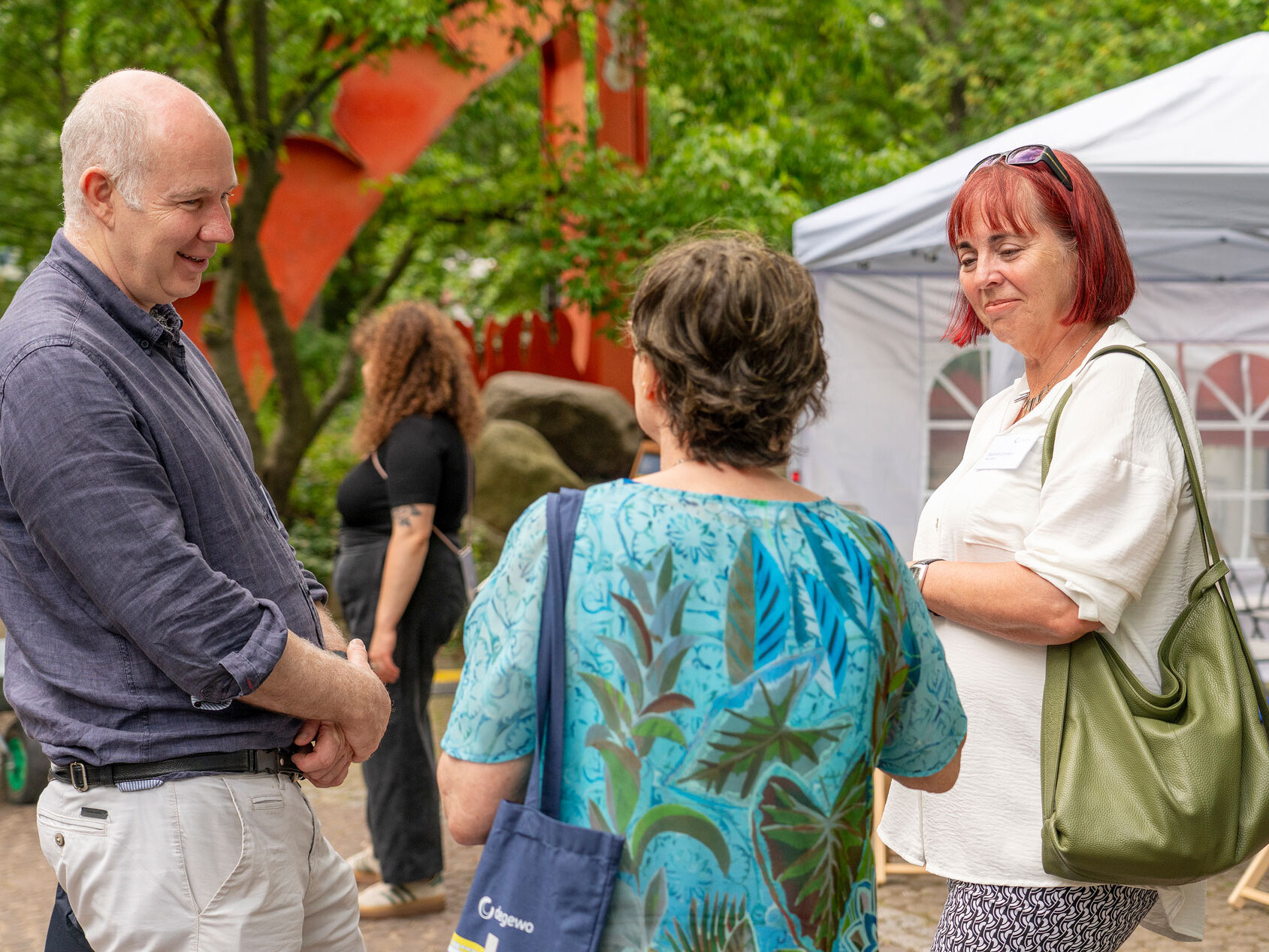 Drei Personen stehen in einem Halbkreis und unterhalten sich. Im Hintergrund Bäume, ein Pavillon und eine weitere Person.