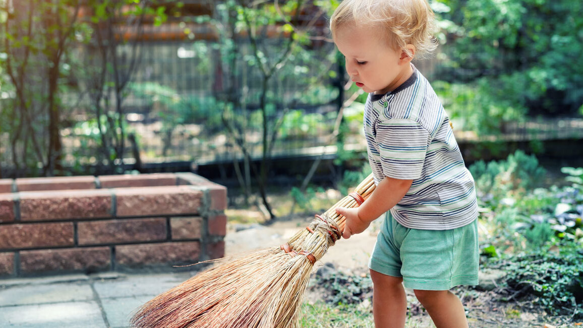 Kleinkind steht im Freien auf einem gepflasterten Weg und hält einen Besen, mit dem es Laub und Schmutz zusammenfegt; im Hintergrund sind Pflanzen und ein Gartenbereich zu sehen.