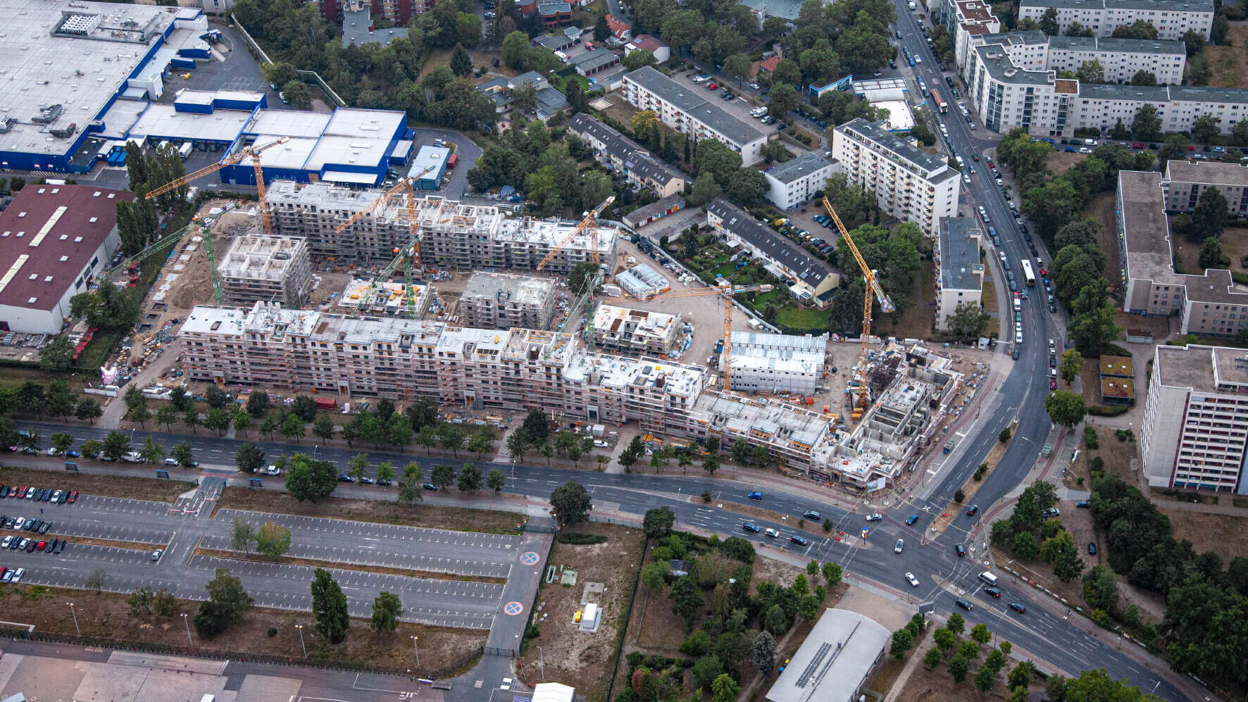 Ein Luftbild zeigt die Baustelle eines Rohbaus mit mehreren Wohnblöcken aus der Vogelperspektive. Dazwischen mehrere Kräne. Um die Baustelle Straßen, weitere Wohnblöcke und Bäume.