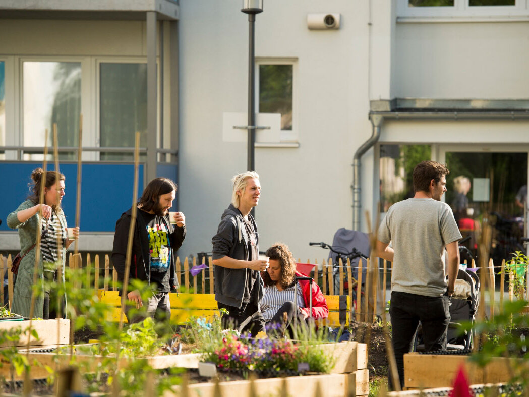 Personen halten sich im Gemeinschaftsgarten auf. Im Hintergrund ist ein Wohnhaus zu sehen.