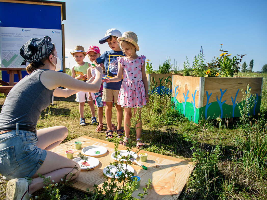 Mehrere Kinder stehen bei einer Kreativaktion im Freien. Eine Betreuerin erklärt eine Bastel- oder Malaufgabe, Farben und Materialien liegen bereit.