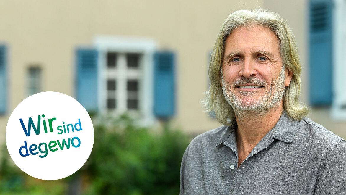 Ein Mann mit grauen Haaren und Bart steht lächelnd im Freien vor einem Gebäude während das degewo Logo mit der Aufschrift Wir sind degewo eingeblendet ist.