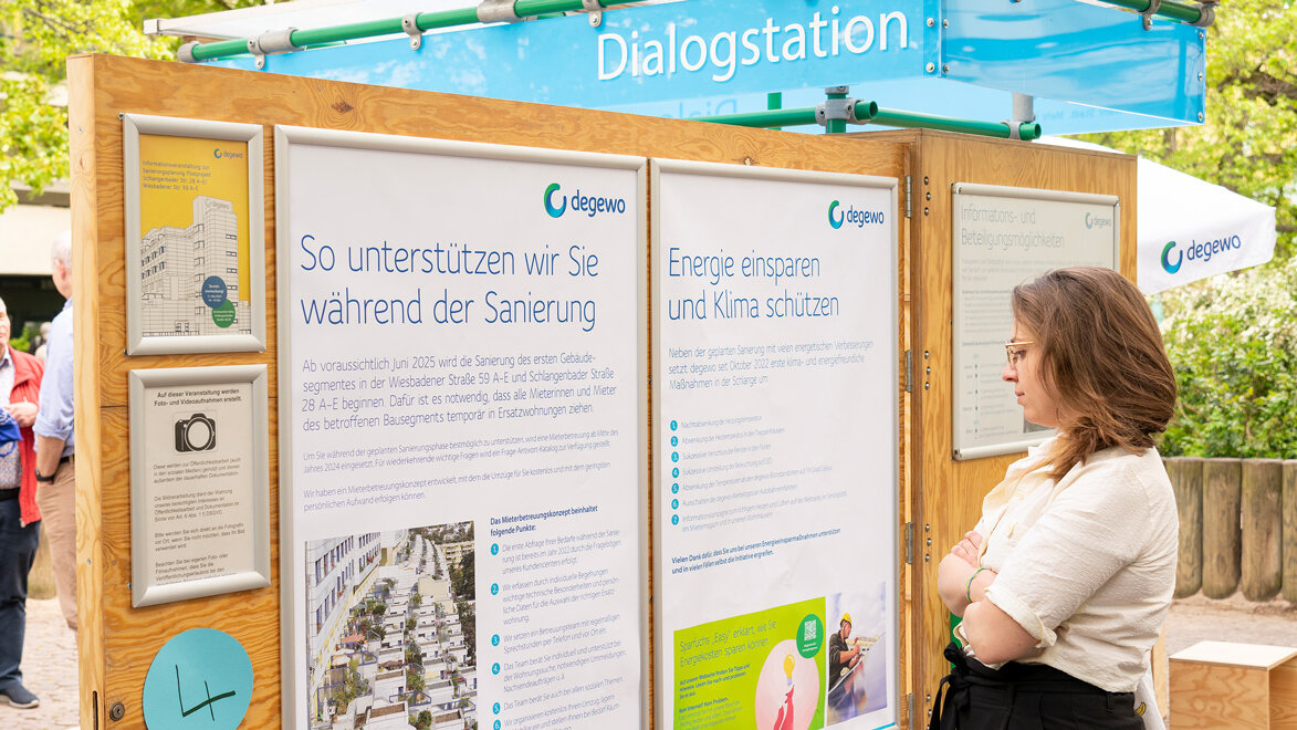 Eine Frau liest Infotafeln an einer Dialogstation im Freien. Hinter ihr stehen weitere Besucher zwischen Bäumen.