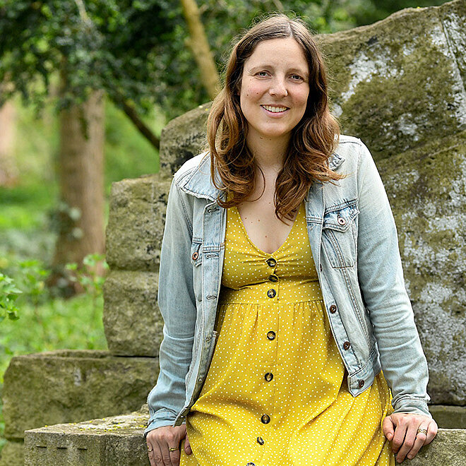 Eine Frau sitzt auf einer Mauer in einem grünen Park. Sie trägt ein gelb gepunktetes Kleid und lächelt in die Kamera.