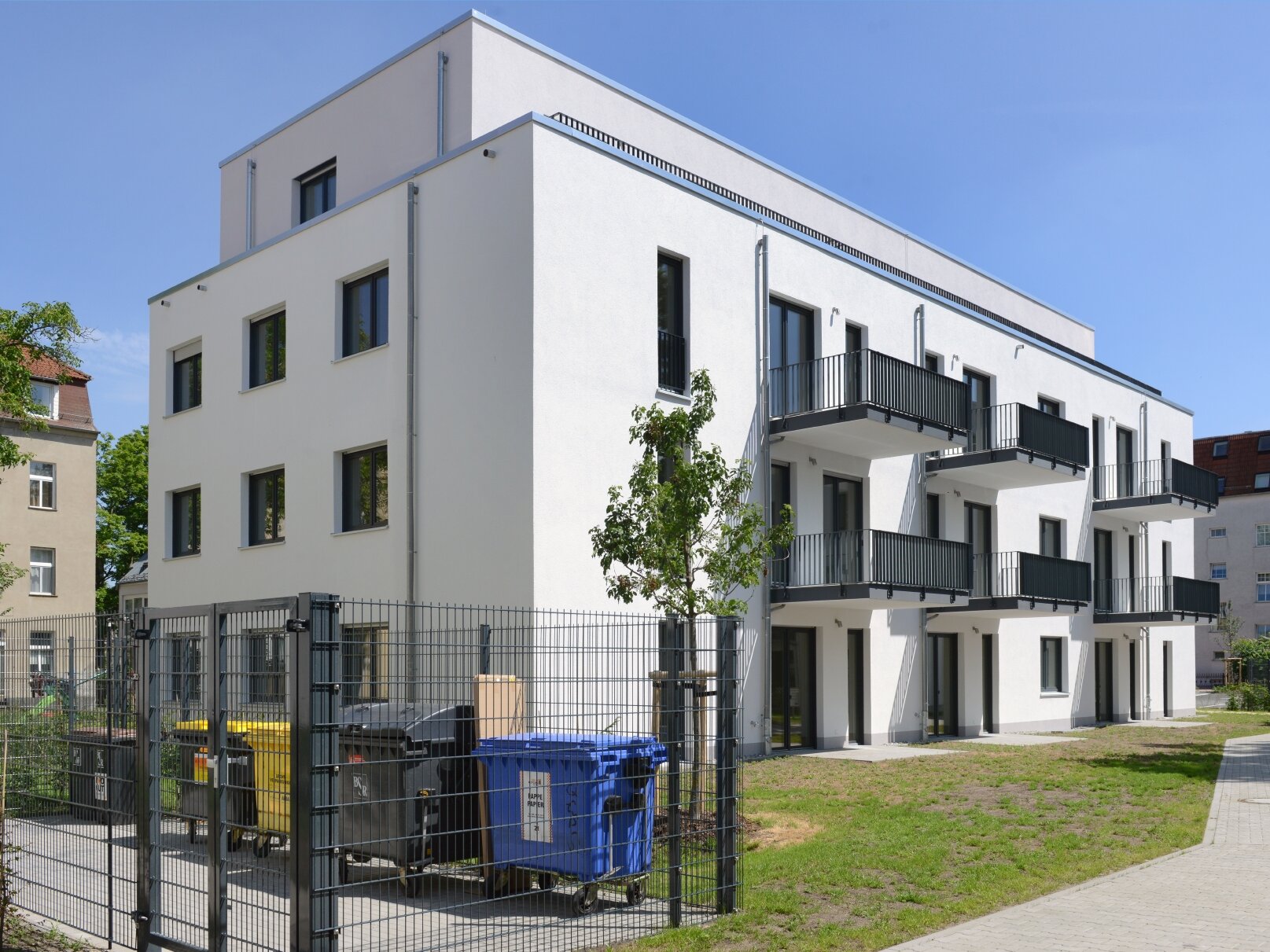 Hofansicht degewo-Neubau Karl-Frank-Straße 33 und 35 mit eingezäunten Mülltonnen, Grünfläche und Weg im Vordergrund.