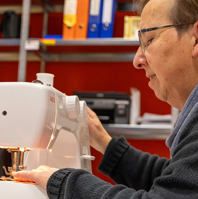 Eine Mitarbeiterin sitzt an der Nähmaschine, im Hintergrund arbeitet ein Mann am Computer.