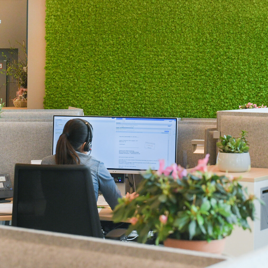 Mitarbeiterin mit Headset arbeitet an einem Computerarbeitsplatz im Kundencenter, moderne Bürofläche mit Trennwänden und begrünter Mooswand im Hintergrund.