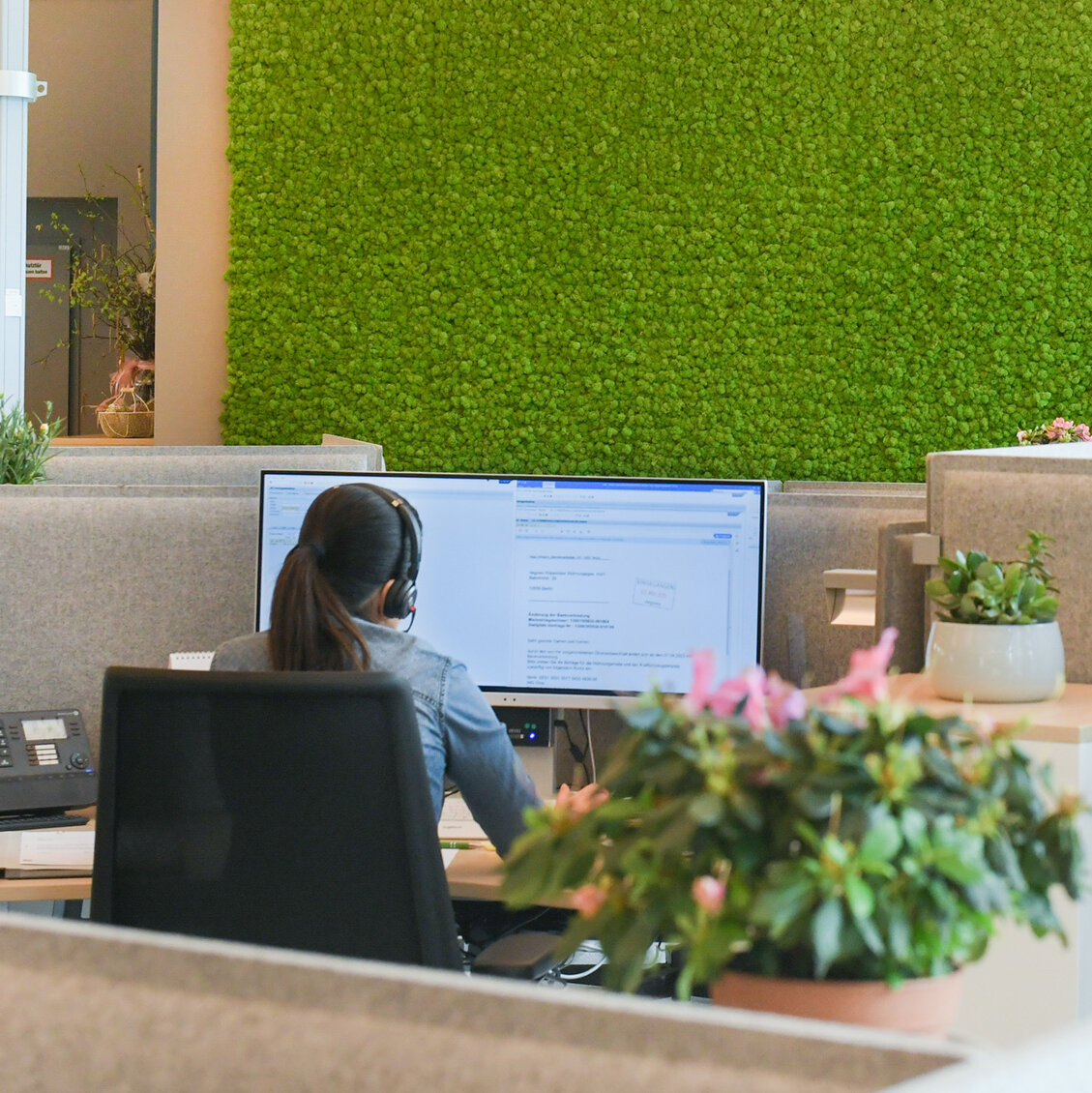 Mitarbeiterin mit Headset arbeitet an einem Computerarbeitsplatz im Kundencenter, moderne Bürofläche mit Trennwänden und begrünter Mooswand im Hintergrund.