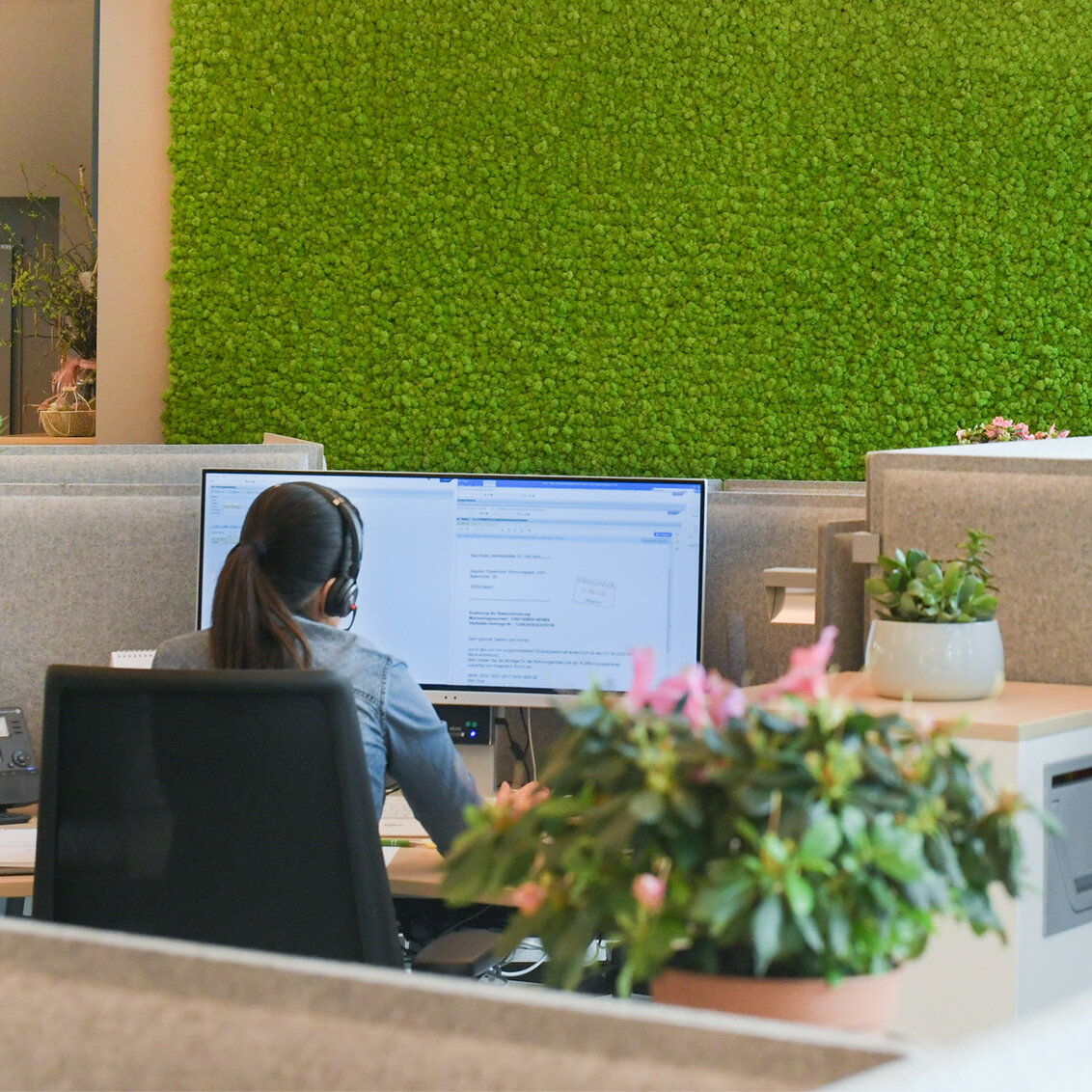 Mitarbeiterin mit Headset arbeitet an einem Computerarbeitsplatz im Kundencenter, moderne Bürofläche mit Trennwänden und begrünter Mooswand im Hintergrund.
