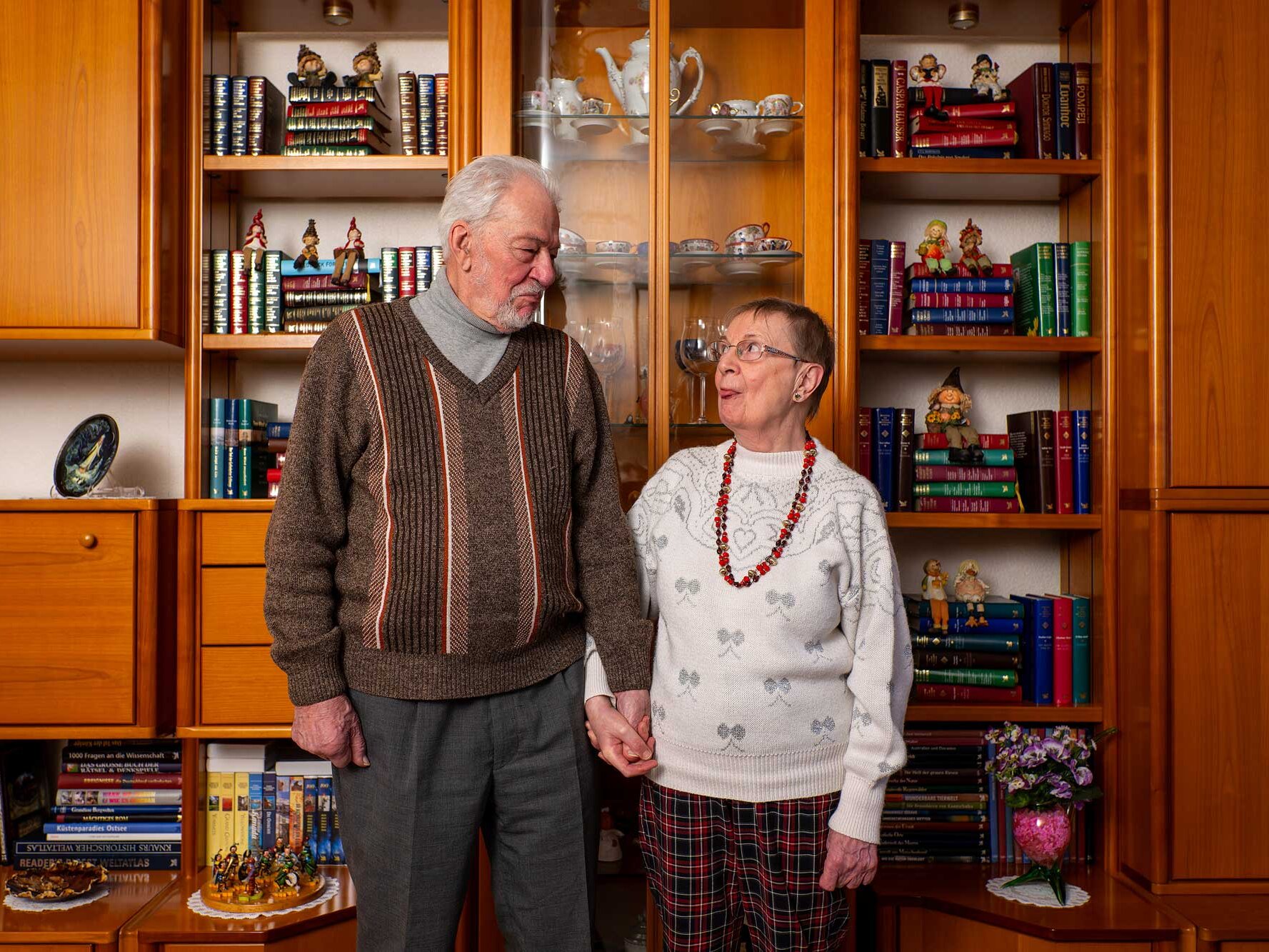 Ein älteres Paar steht Hand in Hand vor einer großen Wohnwand mit Büchern und Porzellan und schaut sich liebevoll an.