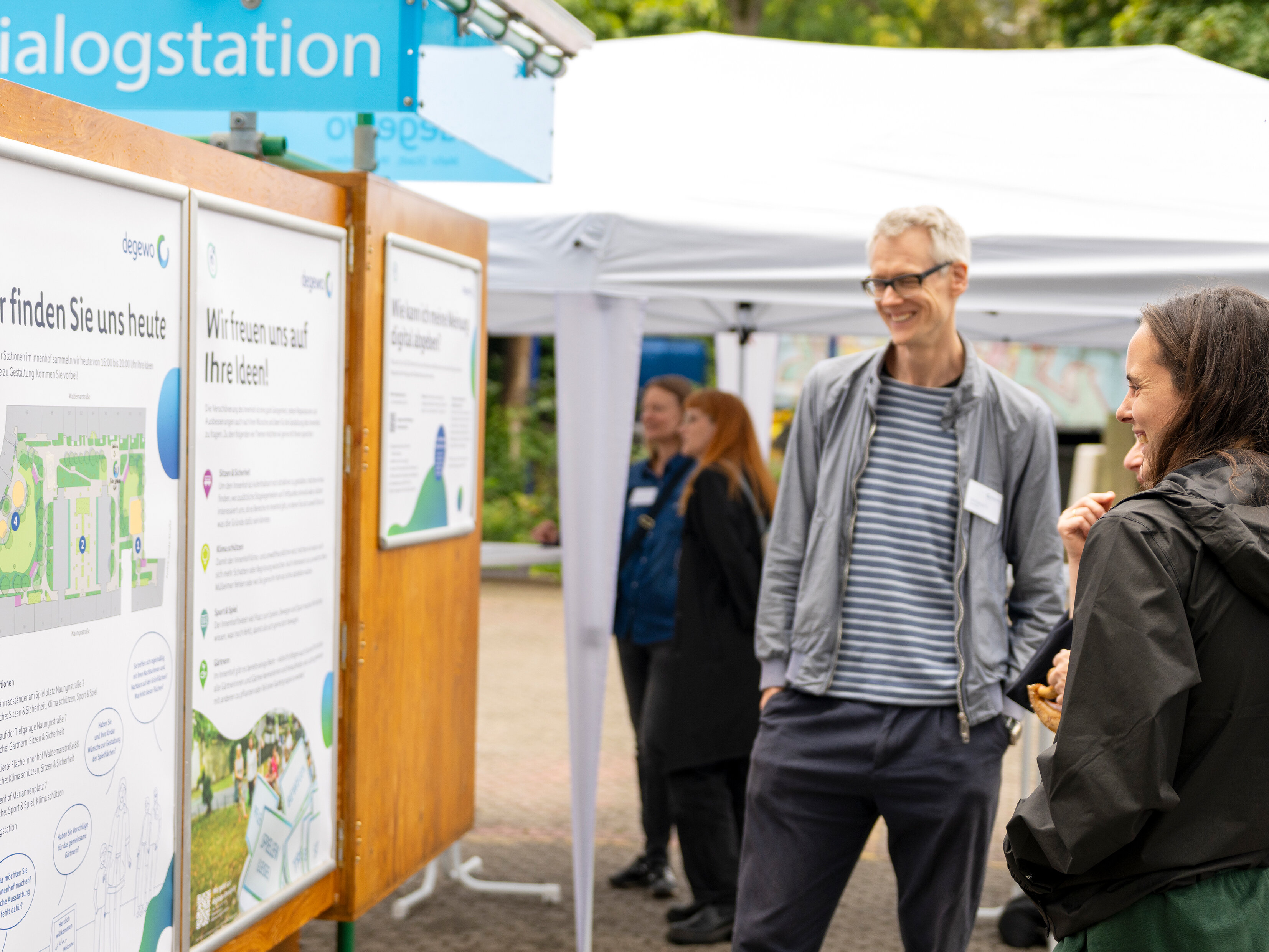Informations- und Dialogstation im Innenhof einer Wohnanlage: Zwei Personen betrachten Projektplakate zur Umgestaltung, daneben ein degewo-Infostand mit Pavillon.