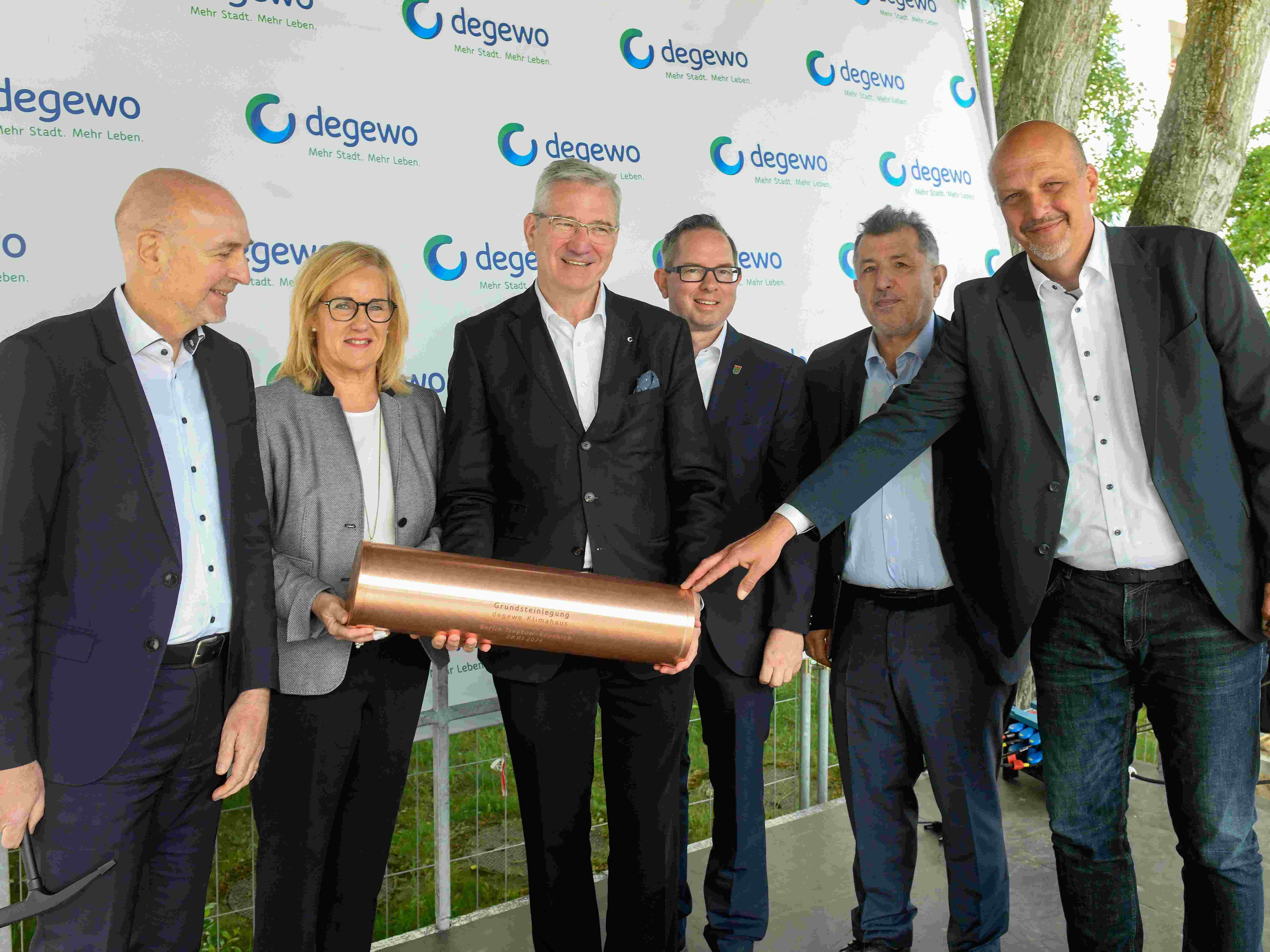 Sechs Personen stehen vor einer degewo-Logowand und halten gemeinsam eine zylindrische Kupferrolle mit Gravur in den Händen. Die Männer und die Frau tragen Businesskleidung mit Jackett bzw. Blazer. Im Hintergrund sind Bäume und ein Bauzaun zu sehen. Die Szene findet im Freien auf einer Bühne statt.