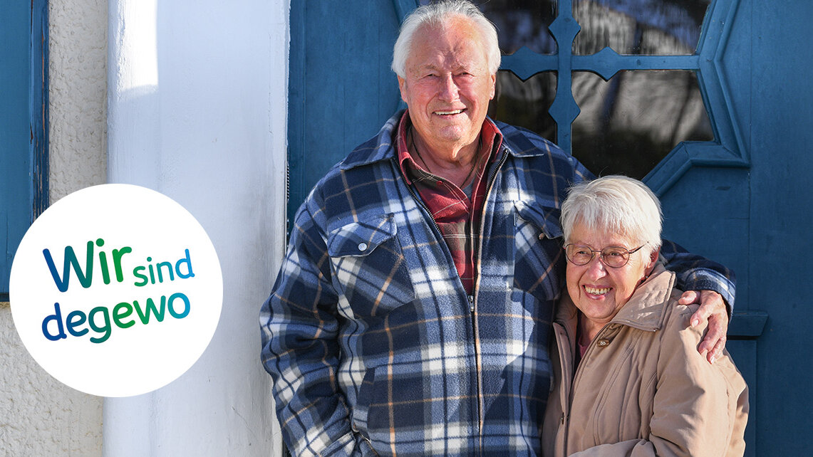 Ein älteres Paar vor der Eingangstür zu ihrem Haus in der Krugpfuhlsiedlung. Der Mann hat den Arm um die Schultern seiner Frau gelegt, die beiden lächeln in die Kamera.