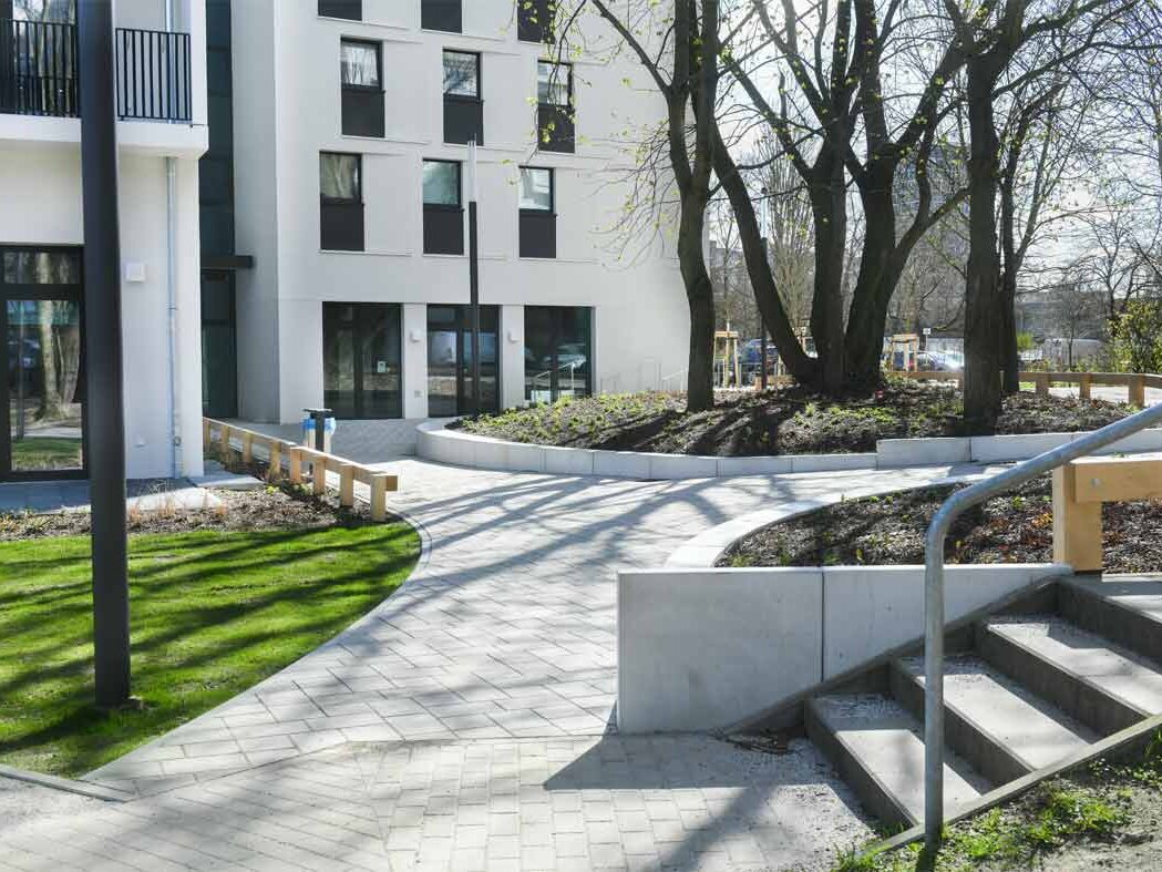 Außenansicht degewo-neubau Friedrich-Kayßler-Weg 1 mit gepflasterten Wegen, Grünflächen und Treppe links im Vordergrund.