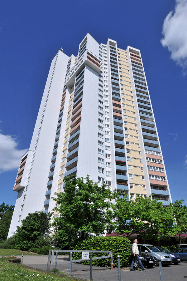 Hohes Wohnhochhaus mit vielen Balkonen vor blauem Himmel, im Vordergrund Bäume, parkende Autos und eine Person auf dem Gehweg.