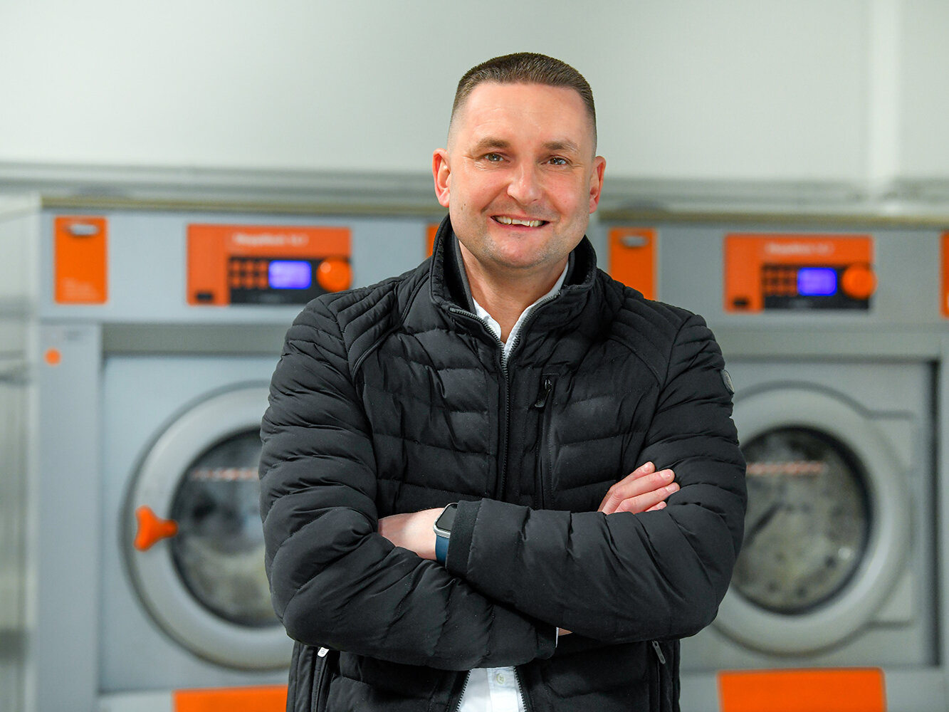 Ein Mitarbeiter steht lächelnd und mit verschränkten Armen vor großen industriellen Waschmaschinen in einem Waschraum.