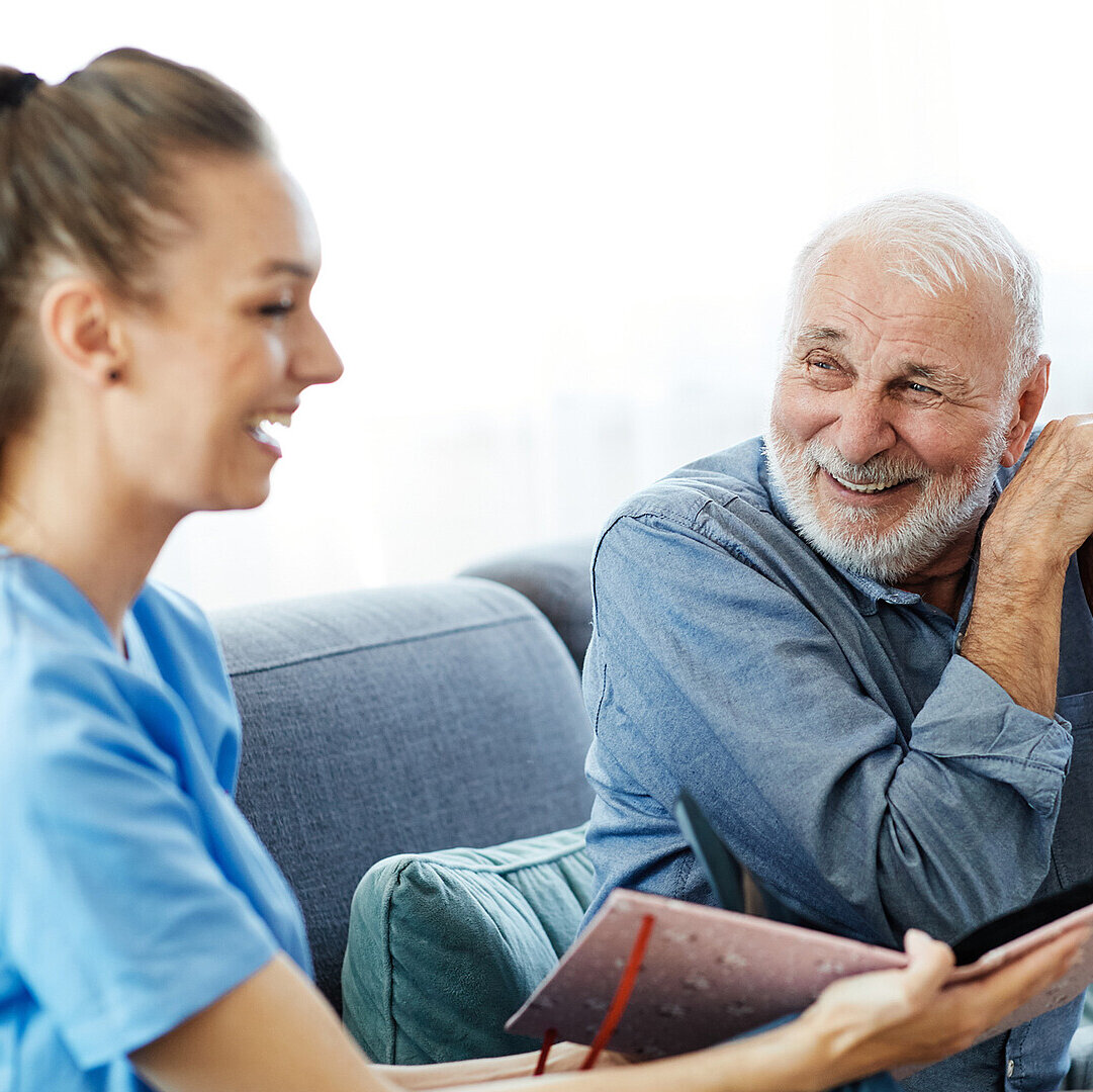 Eine Pflegerin und ein älterer Mann sitzen gemeinsam auf einer Couch und lachen.