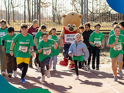Eine Gruppe Kinder rennt auf einem Sportplatz mit dem Maskottchen Berlino.
