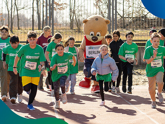 Eine Gruppe Kinder rennt auf einem Sportplatz mit dem Maskottchen Berlino.