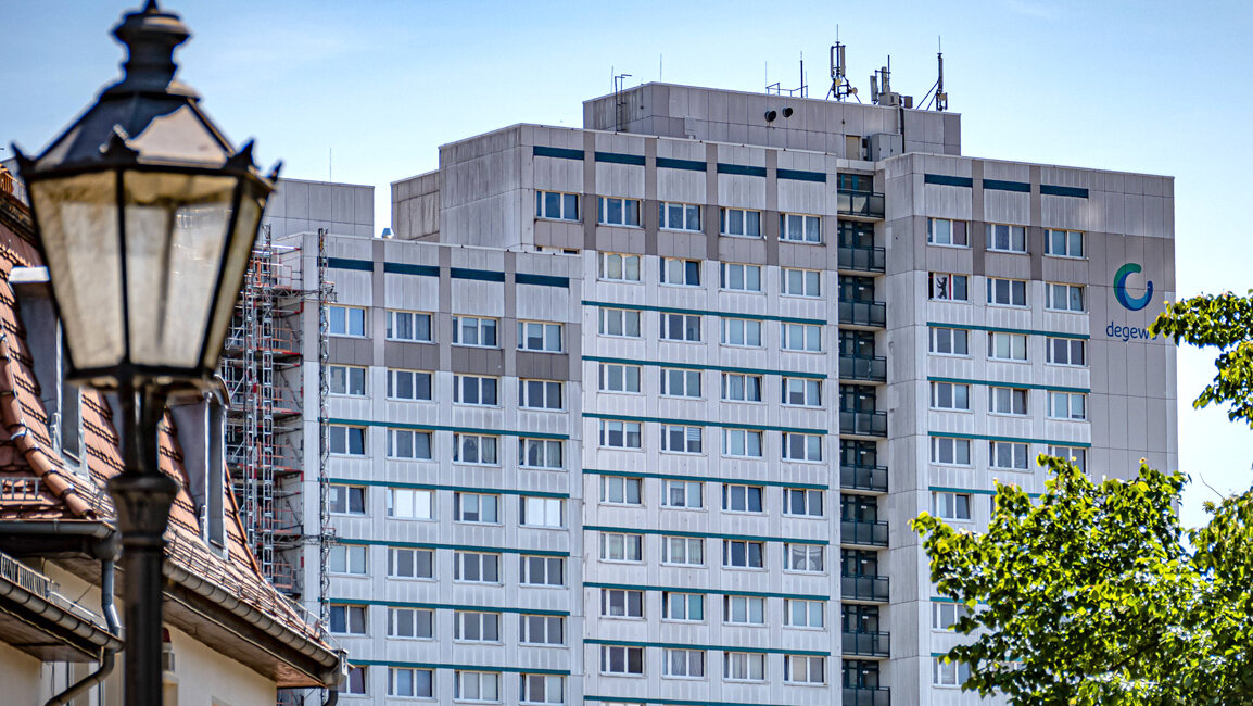 Großes Wohnhochhaus mit vielen Fenstern und dem degewo-Logo an der Fassade, aufgenommen aus der Entfernung; im Vordergrund sind Bäume und ein historischer Laternenmast zu sehen.