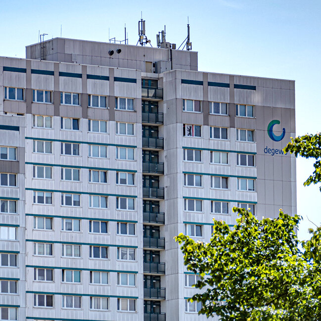 Großes Wohnhochhaus mit vielen Fenstern und dem degewo-Logo an der Fassade, aufgenommen aus der Entfernung; im Vordergrund sind Bäume und ein historischer Laternenmast zu sehen.