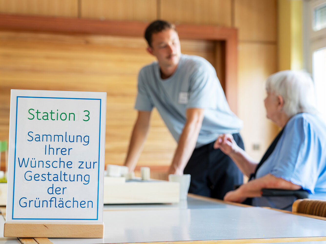 Eine ältere Frau spricht mit einem Mitarbeiter an einem Tisch. Im Vordergrund steht ein Schild mit der Aufschrift „Station 3 – Sammlung Ihrer Wünsche zur Gestaltung der Grünflächen“.