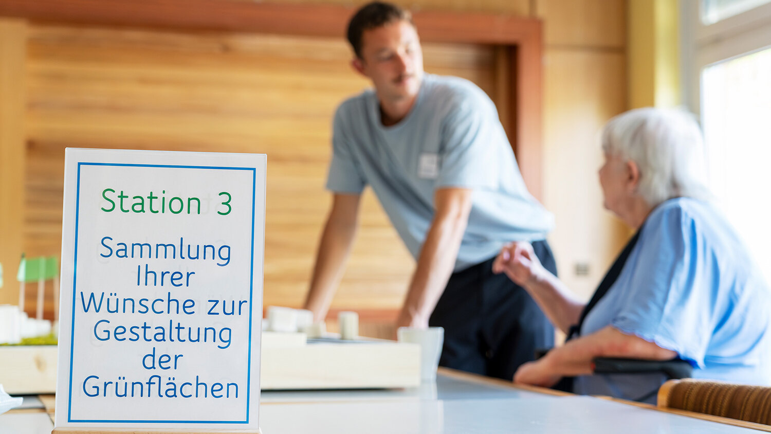 Eine ältere Frau spricht mit einem Mitarbeiter an einem Tisch. Im Vordergrund steht ein Schild mit der Aufschrift „Station 3 – Sammlung Ihrer Wünsche zur Gestaltung der Grünflächen“.