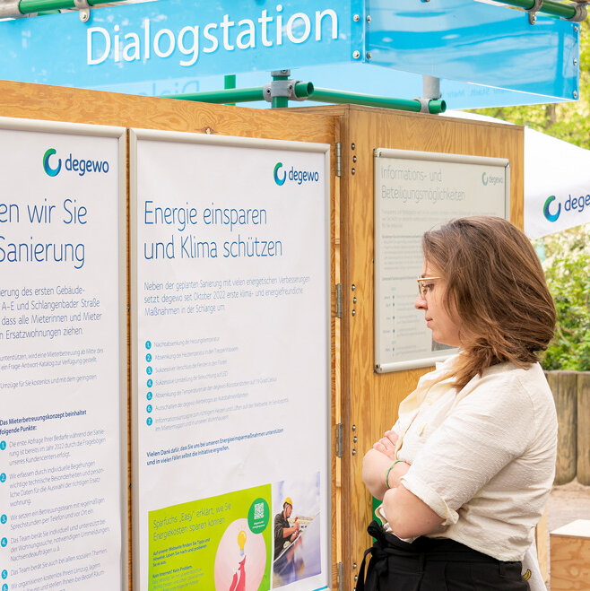 Eine Frau liest Infotafeln an einer Dialogstation im Freien. Hinter ihr stehen weitere Besucher zwischen Bäumen.