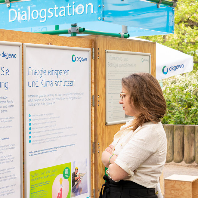 Eine Frau liest Infotafeln an einer Dialogstation im Freien. Hinter ihr stehen weitere Besucher zwischen Bäumen.