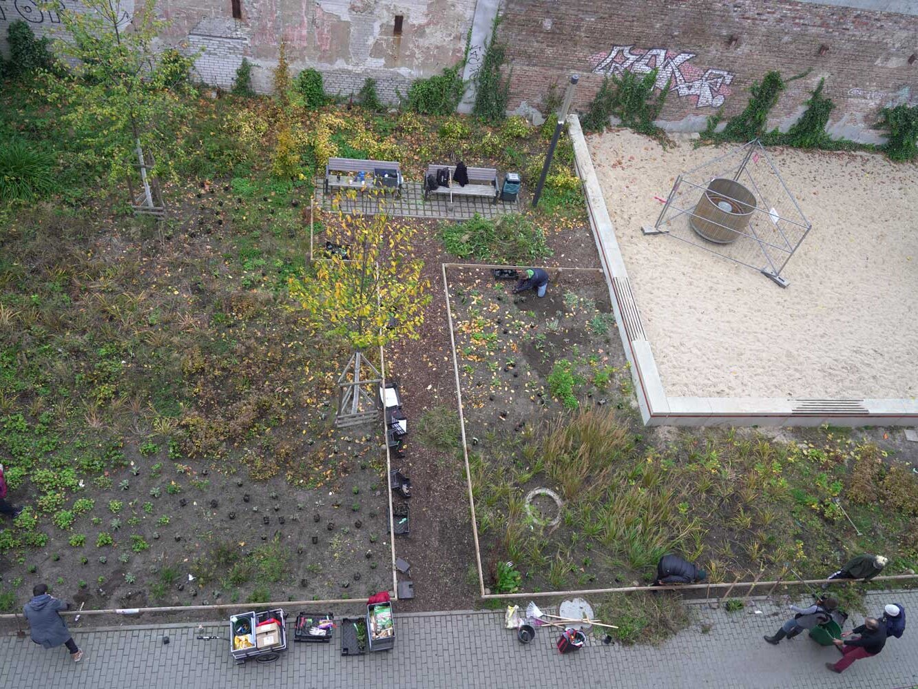 Luftaufnahme einer gepflegten Gartenfläche neben einem Sandspielbereich. Mehrere Personen arbeiten an Beeten und setzen neue Pflanzen.