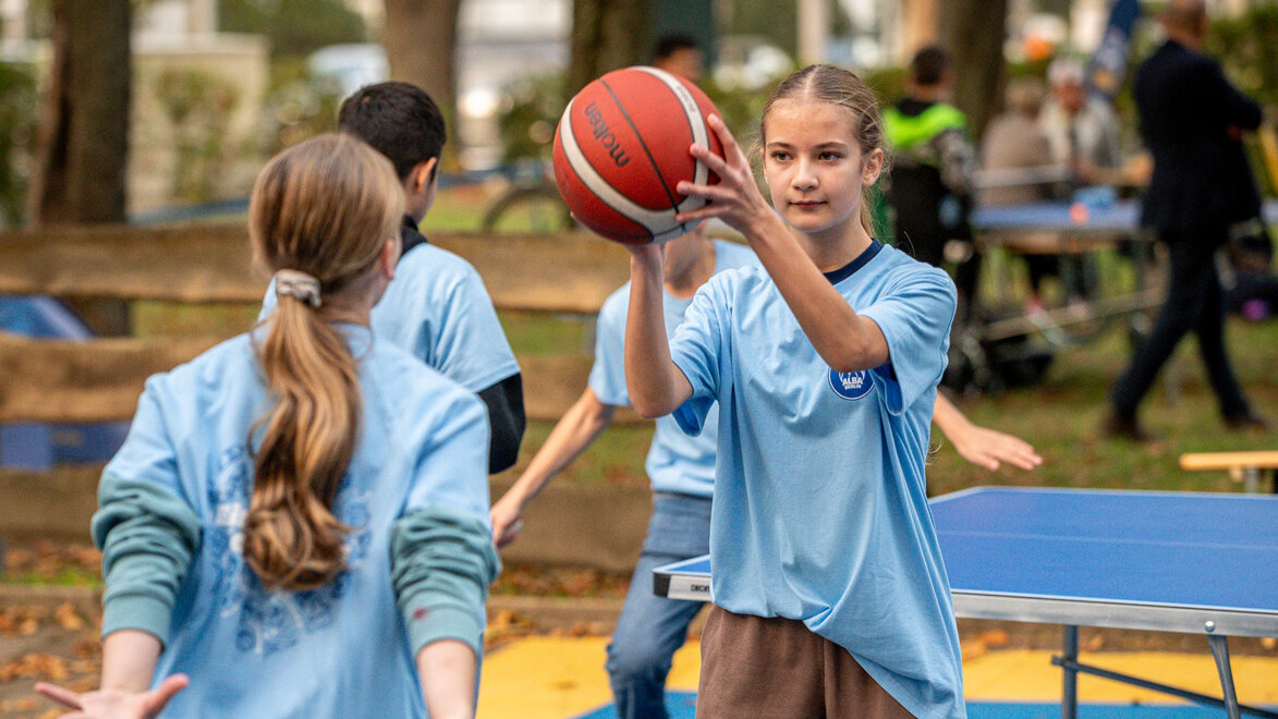 Mädchen in hellblauem Shirt fokussiert beim Basketballspiel, während andere Kinder und Besucher im Hintergrund aktiv sind.
