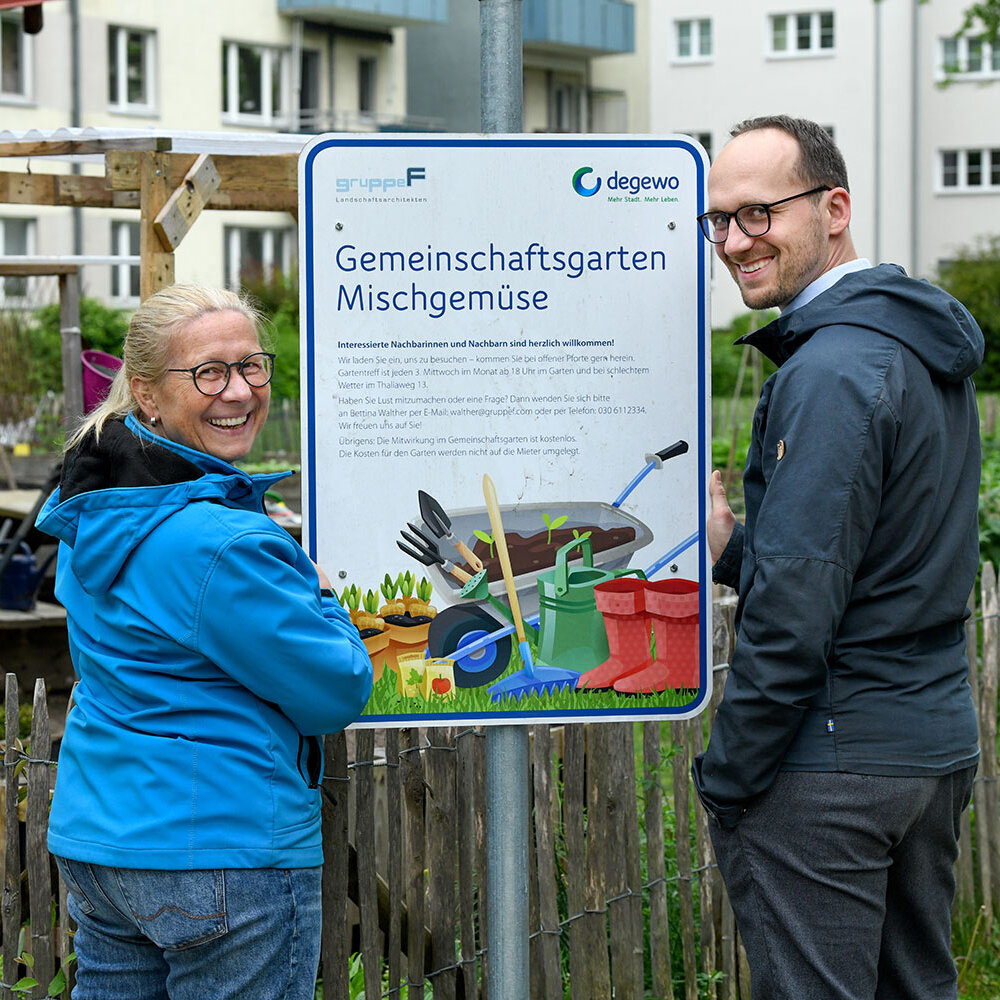 Zwei Mieter stehen vor einem Schild, auf dem Gemeinschaftsgarten Mischgemüse steht. Im Hintergrund ist ein Wohnhaus zu sehen.