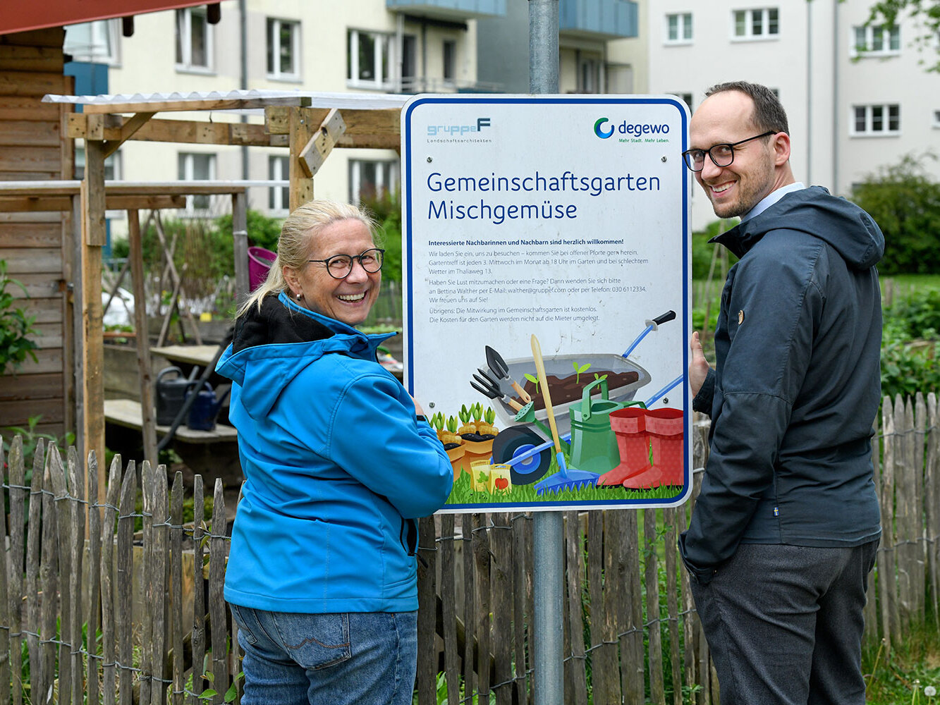 Zwei Mieter stehen vor einem Schild, auf dem Gemeinschaftsgarten Mischgemüse steht. Im Hintergrund ist ein Wohnhaus zu sehen.